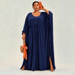 May include: A floor-length navy blue kaftan dress featuring a round neckline, loose flowing sleeves, and a drawstring waist. The garment includes vertical ruffled trim along the side edges and a side slit. A woven tan handbag with an orange ribbon is held.