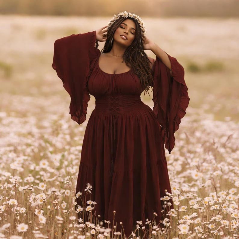 May include: A deep burgundy midi dress featuring a scoop neckline, long flared bell sleeves, and a corset-style lace-up waist detail. The dress has a gathered bodice and a tiered skirt, paired with a white floral crown accessory.