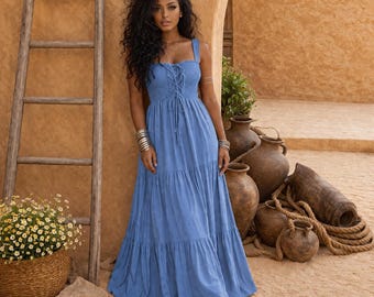 May include: A floor-length blue dress featuring a smocked bodice with a lace-up front detail, wide shoulder straps, and a tiered skirt. The garment has a fitted top section that transitions into a loose, flowing skirt that reaches the floor.