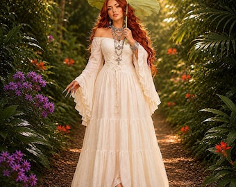 Op de afbeelding: Een off-shoulder crème-kleurige maxi-jurk met lange uitlopende mouwen, een korset met vetersluiting en een gelaagde rok. De jurk wordt gecombineerd met een zilveren gelaagde ketting met kruishangers en een groot groen blad dat als parasol wordt vastgehouden.