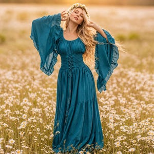 May include: A floor-length teal dress featuring long, wide bell sleeves with ruffled edges, a scoop neckline, and a corset-style lace-up bodice with a smocked waist. The skirt is tiered and flows downward, complemented by a floral crown accessory.