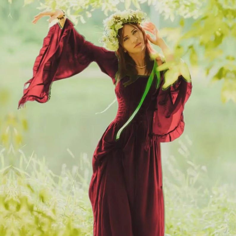 May include: A floor-length deep burgundy dress featuring long, wide bell sleeves with ruffled edges and a fitted bodice. The garment is accented by a bright green ribbon tied at the waist and is paired with a crown of white flowers worn on the head.