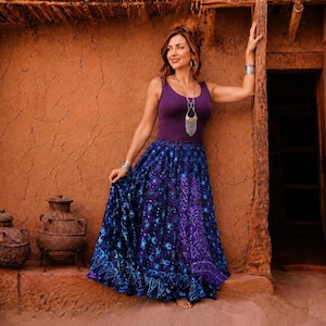 Puede incluir: Una mujer con un top morado y una falda larga y fluida con un estampado azul y morado. El fondo muestra una pared marrón rojiza texturizada y elementos arquitectónicos rústicos.