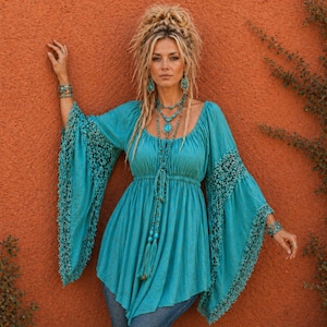 May include: A turquoise peasant top with wide, bell sleeves trimmed with lace. The top has a drawstring neckline and a gathered waist. The model is also wearing turquoise jewellery, including a necklace and earrings. The background is a solid orange-red.
