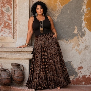 May include: A woman wearing a black tank top and a long, flowing brown skirt with a detailed floral pattern. The skirt has pockets. She is wearing gold jewelry, including a necklace and bracelets. The background features a weathered wall and stone structure.