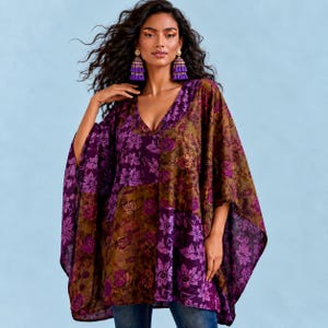 May include: A loose-fitting poncho top featuring a vibrant floral pattern in shades of deep purple, violet, and olive green. The garment has a V-neckline and wide, flowing sleeves. It is paired with long, dangling purple tassel earrings.