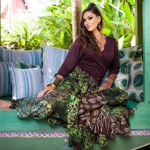 Puede incluir: Una mujer con un top envolvente morado oscuro y una falda larga con un estampado batik verde y marrón. La falda tiene un diseño fluido y en capas. Está sentada en un banco verde con cojines estampados.