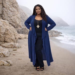 Può includere: Una donna indossa un lungo cardigan blu navy sopra un top e pantaloni neri, in piedi su una spiaggia sabbiosa. Indossa anche una collana con pendente a croce dorata e sandali neri. Lo sfondo presenta rocce, l'oceano e un cielo nebbioso.