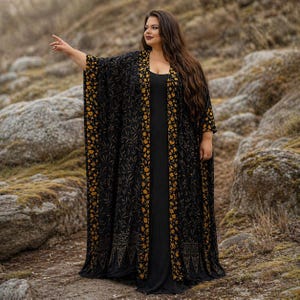 May include: A long black kimono-style robe with a floral pattern in gold and yellow. The robe is worn over a black dress. The background features rocks and a natural setting.