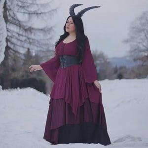 Könnte beinhalten: Ein bordeauxfarbenes Kleid mit langen, ausgestellten Ärmeln und einem mehrschichtigen Rock. Ein schwarzes Korsett schnürt die Taille. Die Person trägt einen schwarzen Hörner-Kopfschmuck. Der Hintergrund ist eine verschneite Landschaft.