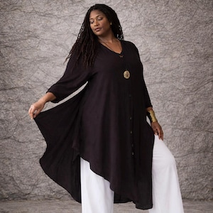 Puede incluir: Una túnica negra con cuello en V y botones. La parte superior tiene mangas tres cuartos y un dobladillo asimétrico. Se combina con pantalones blancos de pierna ancha. Se ve un collar con colgante y una pulsera dorados.