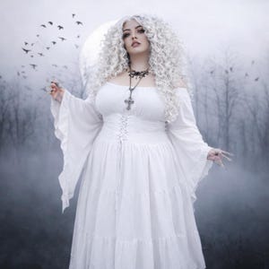 May include: A white off-the-shoulder dress featuring long flared sleeves, a corset-style lace-up bodice, and a tiered skirt. The garment is paired with a silver-toned cross pendant necklace on a delicate chain, worn against a misty, dark forest background.