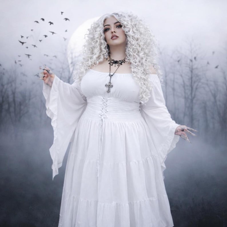 May include: A white off-the-shoulder dress featuring long flared sleeves, a corset-style lace-up bodice, and a tiered skirt. The garment is paired with a silver-toned cross pendant necklace on a delicate chain, worn against a misty, dark forest background.