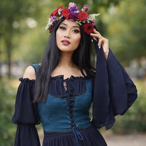 May include: A woman wearing a floral crown of red and purple roses and other flowers. She is wearing a dark teal and navy blue off-the-shoulder dress with a lace-up bodice and long, flowing sleeves. The dress has a Renaissance or medieval style.