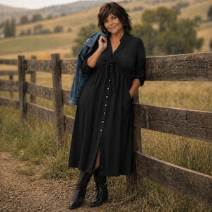 May include: A woman wearing a black button-down midi dress with a tie-front detail and pockets, paired with black boots. A denim jacket is draped over her shoulder. She is leaning against a wooden fence in a field.