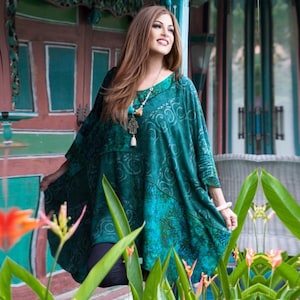 Peut inclure: Une tunique vert turquoise avec un motif. La tunique a une encolure dégagée et des manches trois-quarts. Un long collier avec un pendentif est porté avec la tunique. L'arrière-plan comprend des fleurs et un bâtiment aux accents turquoise et rouges.