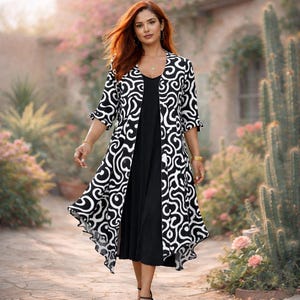 May include: A woman wearing a black dress with a long, open, black and white patterned jacket. The jacket has a swirling design. The dress is knee-length. The woman is walking on a stone path in a garden setting.