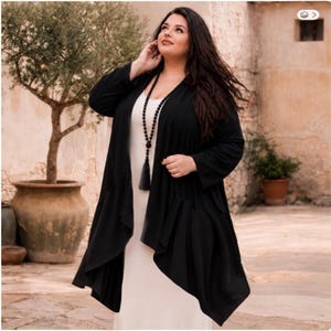 May include: A woman wearing a long black open front cardigan over a white dress. A long black beaded necklace with a tassel is worn. The woman is standing outside near a large potted tree and a stone wall.