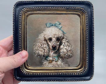 May include: A framed portrait of a poodle with curly white fur, adorned with a blue bow and collar. The painting is set within a black and gold ornate frame, creating a vintage aesthetic.