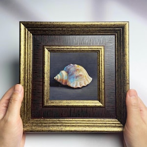 Könnte beinhalten: Ein gerahmtes Kunstwerk mit einer Muschel. Die Muschel ist mit einer Mischung aus Farben bemalt, darunter Blau, Gelb und Orange. Der Rahmen ist eine Kombination aus Gold und Dunkelbraun und verleiht dem Stück einen Hauch von Eleganz.