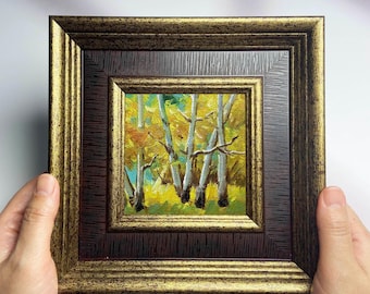 Op de afbeelding: Een kleine, ingelijste schildering van een boslandschap. Het kunstwerk toont witte berken met gele en oranje bladeren, tegen een blauwgroene achtergrond. De lijst is donkerbruin met gouden accenten.