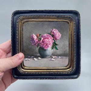 以下が含まれることがあります： 額装された花の静物画。小さな花瓶に入ったピンク色の牡丹が描かれています。フレームは黒色で、内側の縁は金色です。背景はグレーです。油絵です。