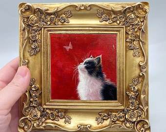 May include: A framed painting of a cat looking up at a butterfly. The cat has black and white fur and is set against a red background. The ornate gold frame features floral detailing.