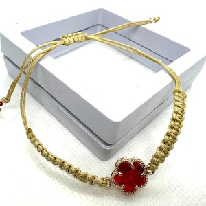 Op de afbeelding: Een goudkleurige verstelbare armband met een rode bloemvormige bedel. De bedel is bezet met kleine, heldere steentjes. De armband is gemaakt van gevlochten koord en wordt gepresenteerd op een wit oppervlak.