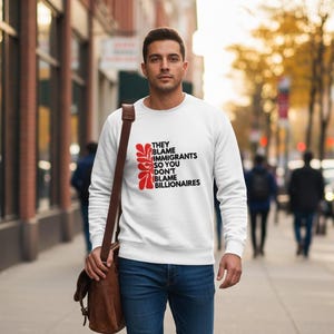 May include: White sweatshirt with the text "THEY BLAME IMMIGRANTS SO YOU DON'T BLAME BILLIONAIRES" in black and red. The sweatshirt has a crew neck and long sleeves. The person is wearing blue jeans and a brown shoulder bag.