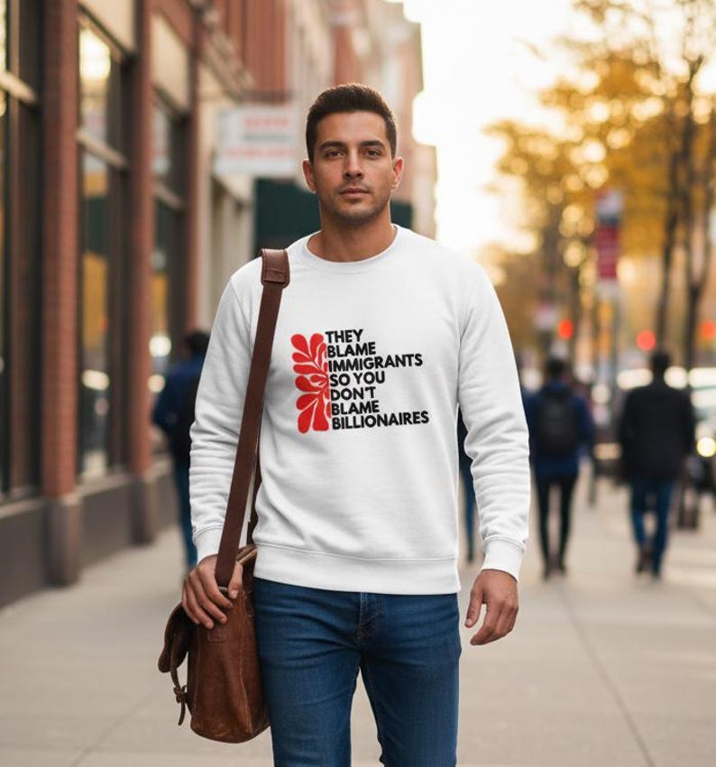 May include: White sweatshirt with the text "THEY BLAME IMMIGRANTS SO YOU DON'T BLAME BILLIONAIRES" in black and red. The sweatshirt has a crew neck and long sleeves. The person is wearing blue jeans and a brown shoulder bag.