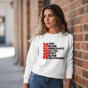 May include: White sweatshirt with the text "THEY BLAME IMMIGRANTS SO YOU DON'T BLAME BILLIONAIRES" in black and red. The sweatshirt has a crew neck and long sleeves. The design includes a red floral graphic.
