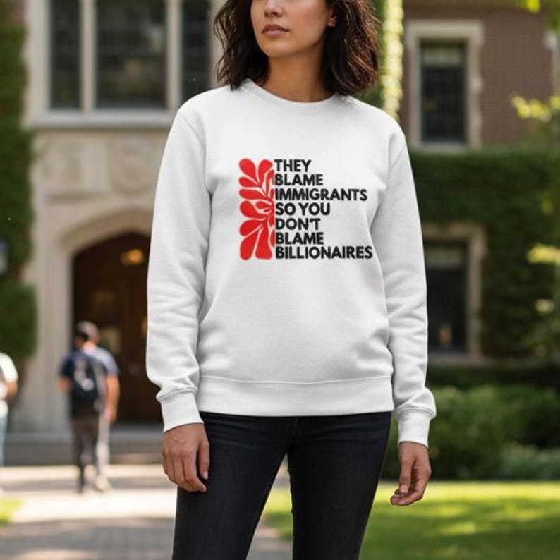 May include: White sweatshirt with the text "THEY BLAME IMMIGRANTS SO YOU DON'T BLAME BILLIONAIRES" in black and red. The sweatshirt has a crew neck and long sleeves. The graphic is on the front of the sweatshirt.