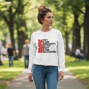 May include: White sweatshirt with the text "THEY BLAME IMMIGRANTS SO YOU DON'T BLAME BILLIONAIRES" in black and red. The sweatshirt is worn by a person in blue jeans, walking on a path in a park.