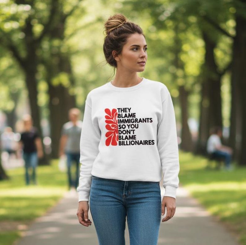 May include: White sweatshirt with the text "THEY BLAME IMMIGRANTS SO YOU DON'T BLAME BILLIONAIRES" in black and red. The sweatshirt is worn by a person in blue jeans, walking on a path in a park.