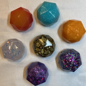 May include: Seven faceted, gem-like objects in various colors. The colors include orange, teal with glitter, orange, lavender with glitter, gold, purple with glitter, and dark purple with glitter. The objects are arranged on a white surface.