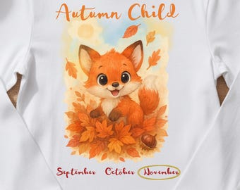 Pode incluir: Uma t-shirt branca de manga comprida com um gráfico de uma raposa de desenho animado sentada em folhas de outono. O texto diz Autumn Child acima da raposa e September, October, November abaixo, com November circulado num oval dourado.