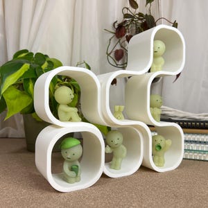 May include: A white, wavy, modular shelf unit with six compartments, each housing a small, green figurine. The shelf is set against a backdrop of plants and a white curtain, creating a minimalist aesthetic.