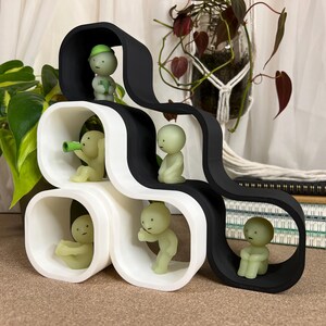 May include: A modern, abstract shelf unit in black and white, featuring rounded cubby holes. Small, pale green figurines are positioned within the cubbies, some holding objects. The shelf is a decorative piece, possibly for displaying small items.