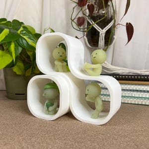 May include: A white, modern sculpture with four rounded cubby holes, each containing a small, light green figurine. The figurines are in various poses. The sculpture is on a brown surface, with plants in the background.