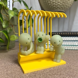 May include: A yellow decorative cage with three small, pale green figures inside. One figure is sitting, another has a pacifier, and the third is standing. The cage has a yellow base and arched top, with vertical bars. A plant and notebooks are in the background.
