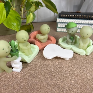 May include: A collection of small, pale green figurines in various poses, some seated on pastel-colored platforms. One figurine holds a small potted cactus. A white, abstract shape is in the center. The figurines have simple, minimalist features.