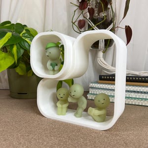 May include: A white, modern shelf unit with rounded corners, holding four small, green-toned figurines. The figurines have simple, minimalist designs. The shelf is on a neutral surface, with plants and notebooks in the background.