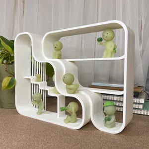 May include: A white, modern, sculptural shelf unit with curved edges and multiple levels. Small, green, humanoid figurines are placed on the shelves, each in a different pose. The shelf is set against a neutral background.