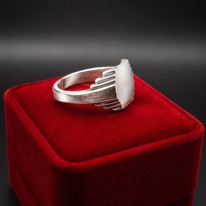 Op de afbeelding: Een zilveren ring met een glad, plat, rechthoekig oppervlak en een getrapt, gelaagd ontwerp op de schouders. De ring heeft een gepolijste metalen afwerking met zichtbare lichtreflecties, wat een gestructureerde, geometrische vormgeving toont.