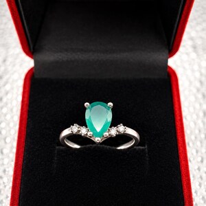 Op de afbeelding: Een zilverkleurige ring met een centrale peervormige groene edelsteen, geflankeerd door twee kleine heldere ronde stenen aan weerszijden van de ringband. De ring wordt gepresenteerd in een met zwart fluweel bekleed juwelendoosje.