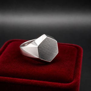 Op de afbeelding: Een zilverkleurige zegelring met een plat, zeshoekig bovenvlak met een geborstelde metalen afwerking. De ringband en de zijkanten van de zeshoekige kop hebben een glad, gepolijst en reflecterend oppervlak dat contrasteert met de textuur bovenop.