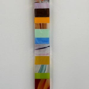 May include: A vertical, rectangular art piece composed of stacked, colorful glass blocks. The blocks display a variety of colors including red, blue, orange, yellow, green, and white. The artwork is mounted against a white wall.
