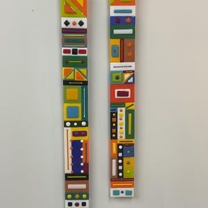 May include: Two vertical, rectangular art pieces with colorful geometric designs. The pieces feature a variety of shapes and colors, including orange, green, yellow, blue, and white. The artwork is mounted on a white background.