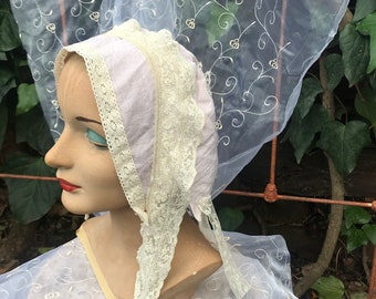 May include: A pale pink bonnet with white lace trim and a sheer veil with embroidered floral designs.