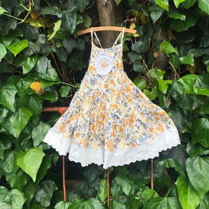 May include: A white sleeveless dress with a yellow and gray floral print and white crochet trim. The dress is hanging on a wooden hanger against a backdrop of green leaves.
